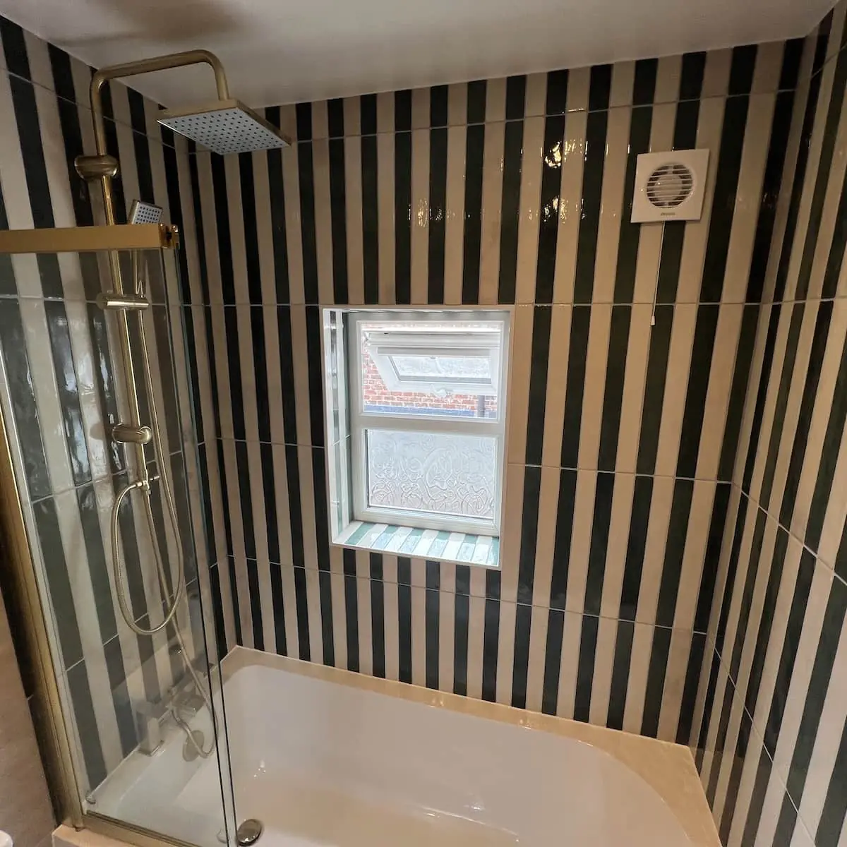 Small Eccles bathroom with vertical green and cream striped tiles, brass-finish shower, and a bath with hinged screen