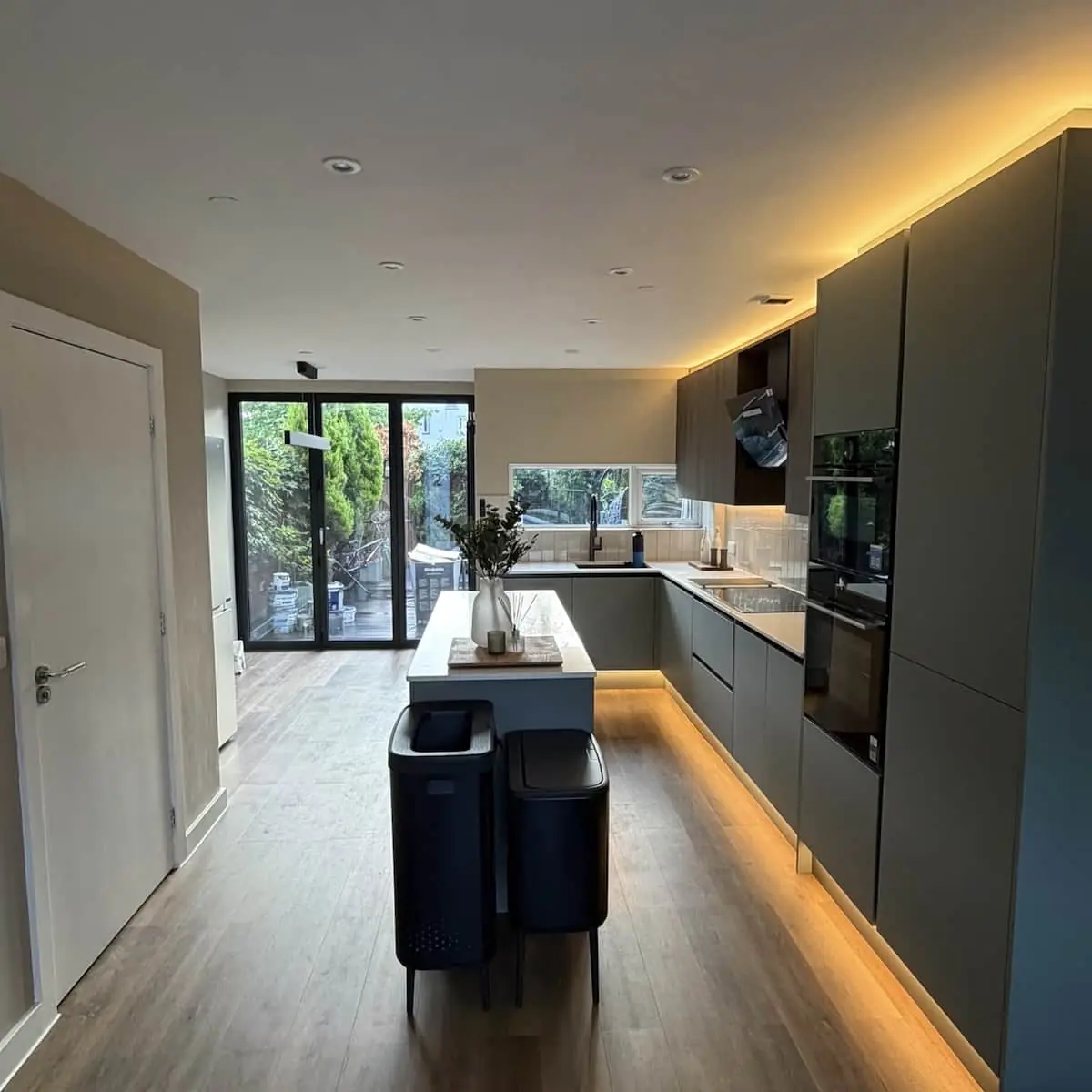 Long handleless kitchen with island, built-in appliances, and glazed doors to the garden, lit by warm LED strips