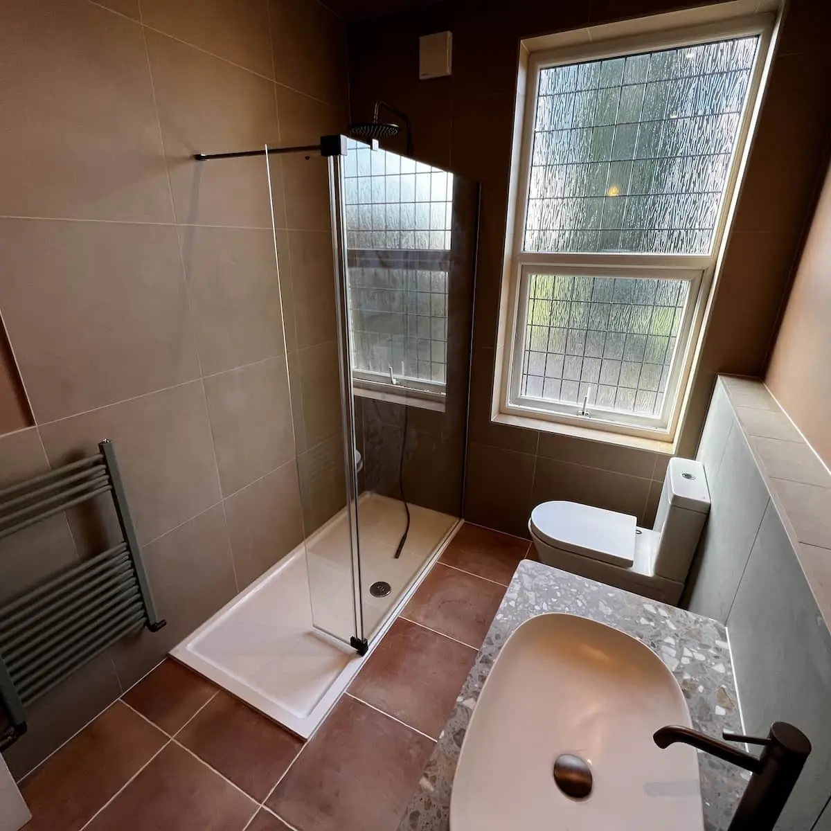 Bury bathroom with terracotta floor, warm brown wall tiles, sage radiator, and terrazzo-style vanity top with oval basin